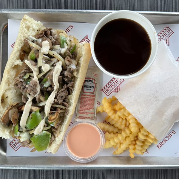 Bruchi's cheesesteak sub with fries, dipping sauce, horseradish packet, and a drink on a tray.