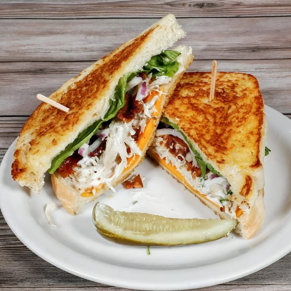A grilled chicken sandwich, cut in half, with cheese, lettuce, onions, and bacon, served with a pickle.