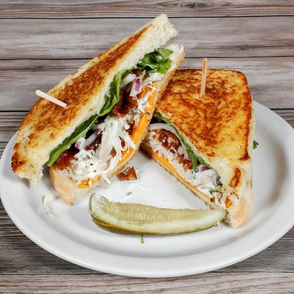 Bruchi's grilled sandwich, cut in half, filled with meat, cheese, lettuce, and onions, served with a pickle.