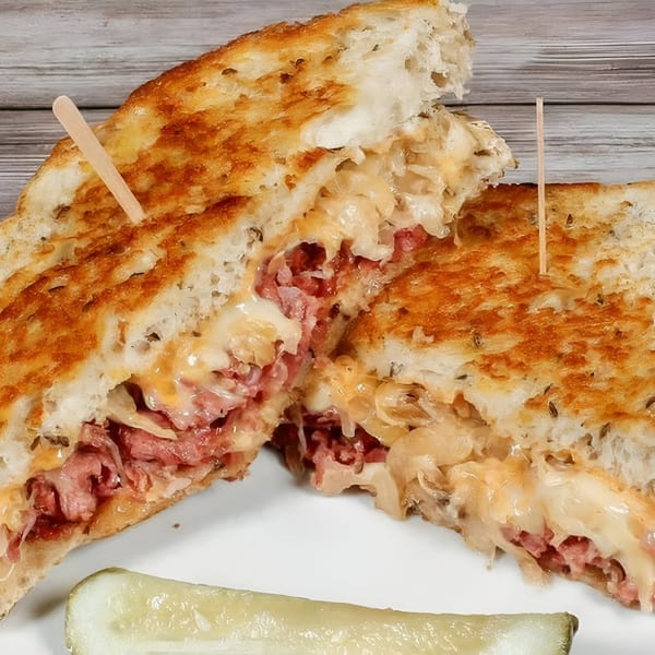 Bruchi's Reuben sandwich, cut in half, revealing corned beef, sauerkraut, and melted cheese, with a pickle spear.