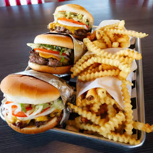 Three Bruchi's cheeseburgers with lettuce, tomato, onion, and cheese, alongside crinkle-cut fries on a tray.