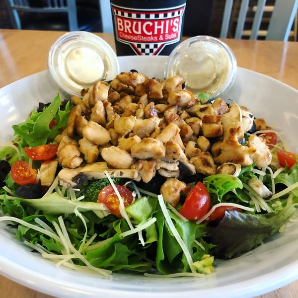 Bruchi's chicken salad with lettuce, tomatoes, olives, and dressing cups on a wooden table.